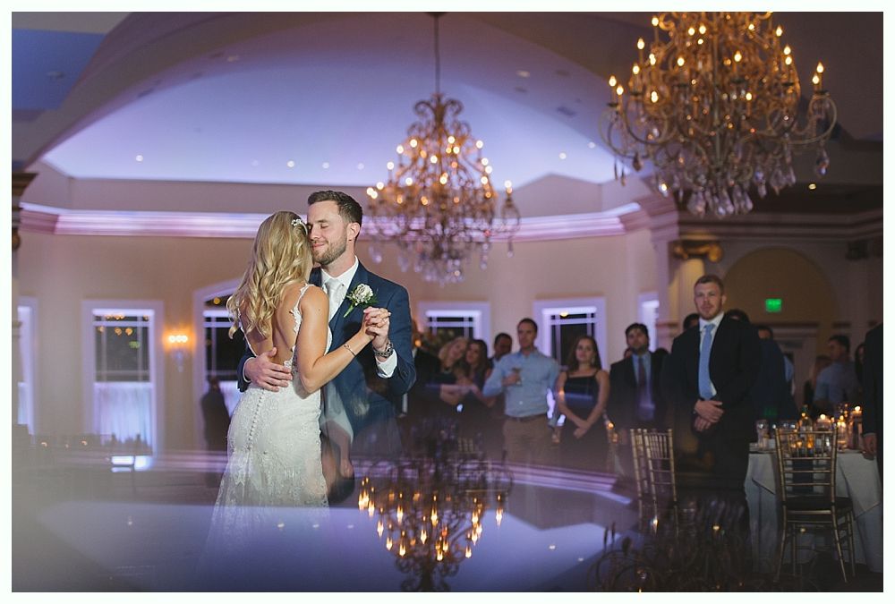 Couple dances at wedding reception; ornate chandeliers, guests watch, reflective dance floor.