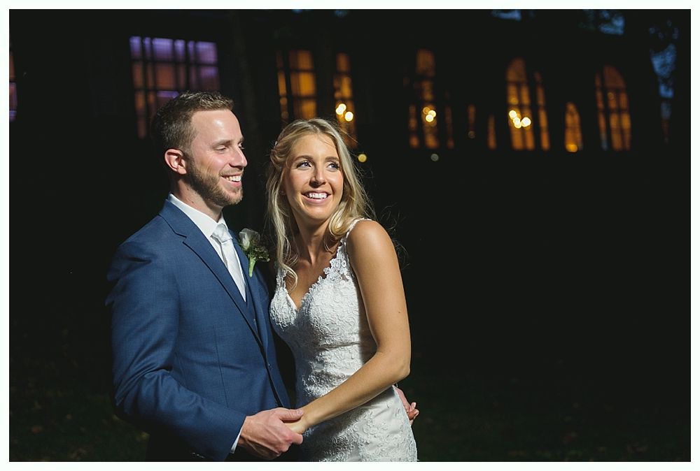 Wedding couple, holding hands, smiling; lit against a dark background with building lights in the distance.