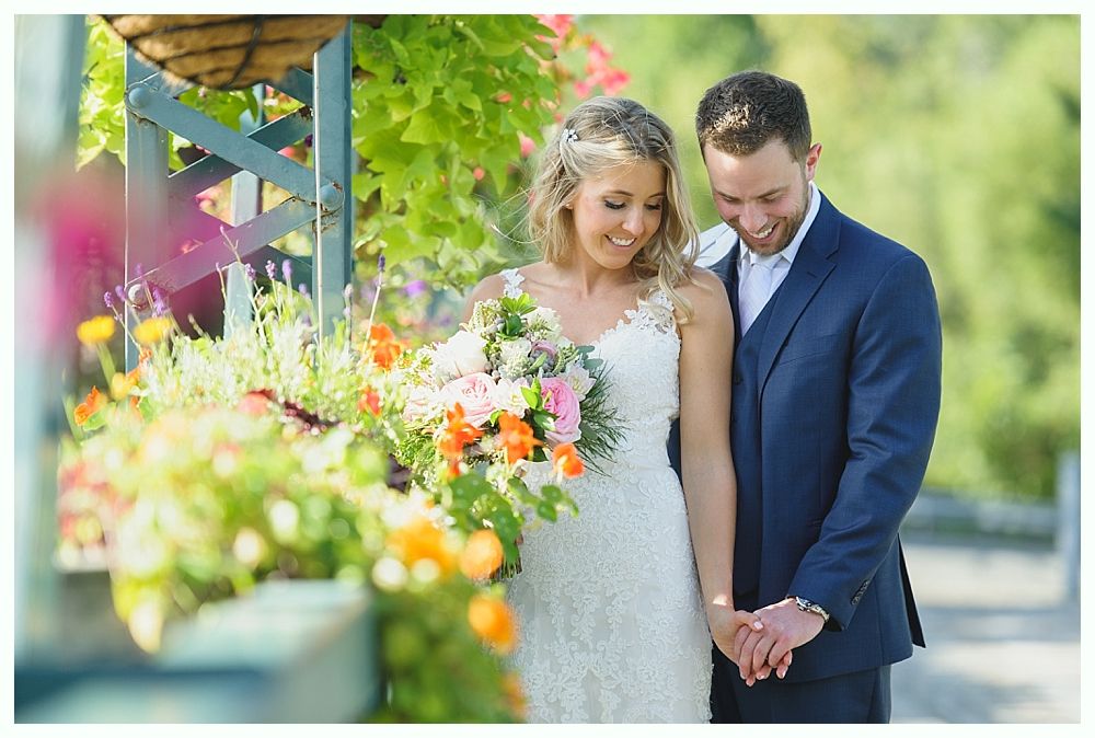 Bride and groom holding hands, smiling. She wears a white lace dress and holds a bouquet, he wears a blue suit. Beside flowers.