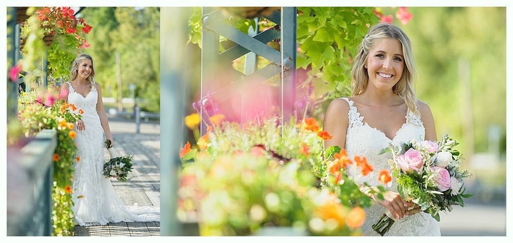 Bride in white dress holding flowers, smiles, stands outdoors by a bridge with colorful flowers.