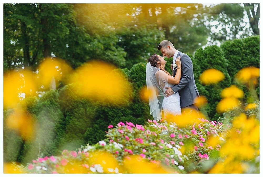 Wedding couple embraces among flowers in a garden. The bride wears a veil; the groom, a suit.