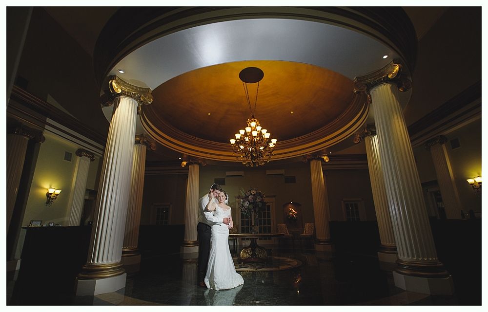 Couple embraces under ornate dome with columns and chandelier.