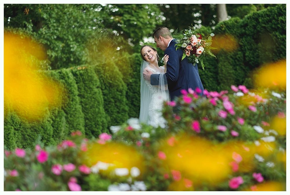 Bride and groom embrace in a garden; bride wears a veil and holds flowers; green hedges and colorful flowers surround them.