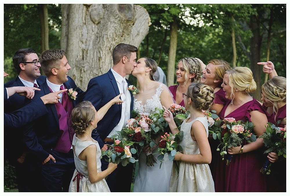 Wedding party with bride and groom kissing, surrounded by bridesmaids and groomsmen, outdoors.