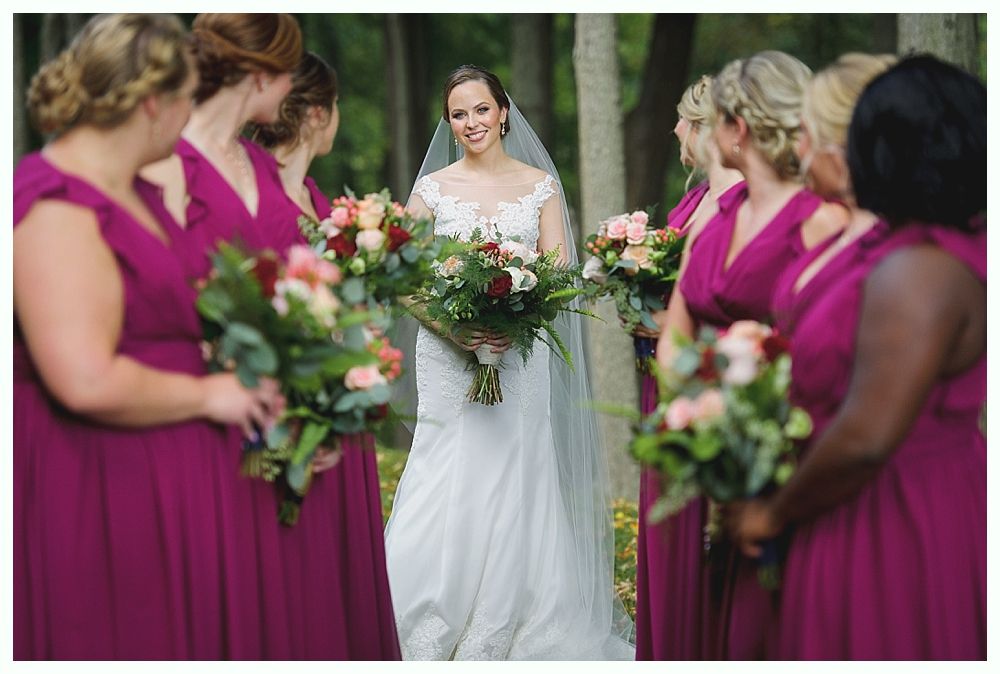 Bride in white gown smiles at bridesmaids in magenta dresses, holding bouquets. Outdoor setting.