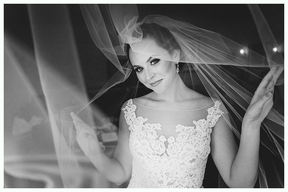 Bride in a wedding dress smiles, veil flowing around her, hands raised slightly.