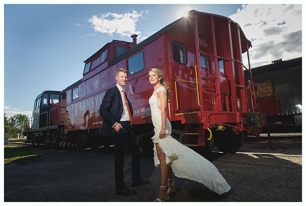 Couple in wedding attire poses next to a red train caboose on a sunny day.