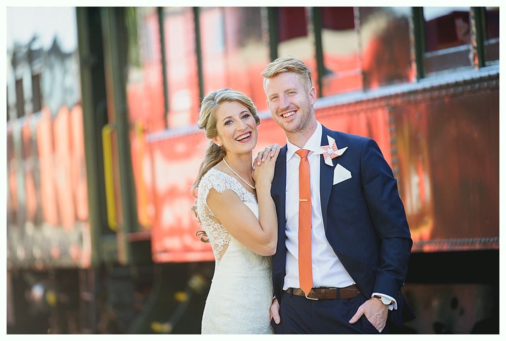 Bride and groom smiling beside a train car. The man wears a navy suit with an orange tie, the woman a white dress.