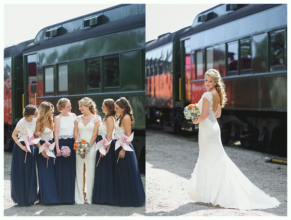 Wedding party posing by a train. Bridesmaids wear navy and white, bride in a white gown. Outdoors.
