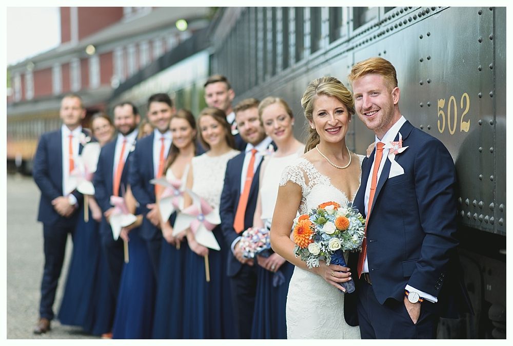 Wedding party in navy and coral leaning against a train; bride and groom smile.
