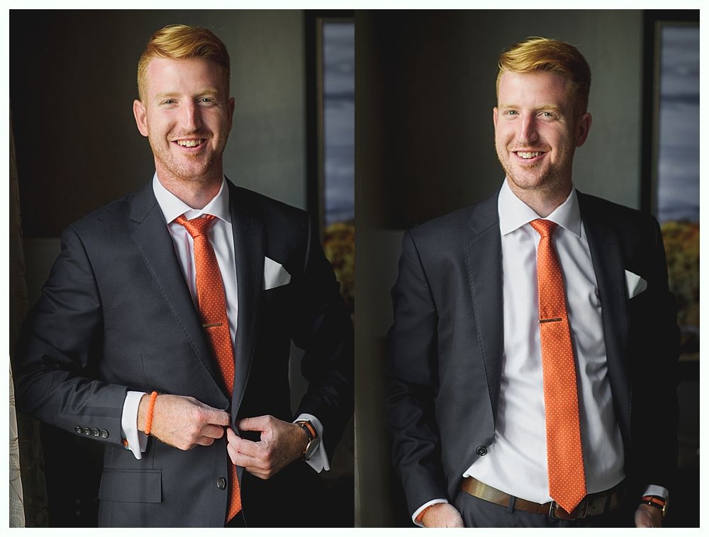 Man in a dark suit with an orange tie, smiling, in front of a window.