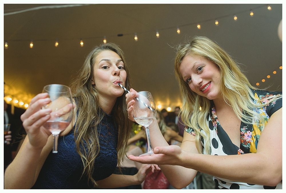 Two women holding wine glasses, one blowing, other gesturing, under string lights.