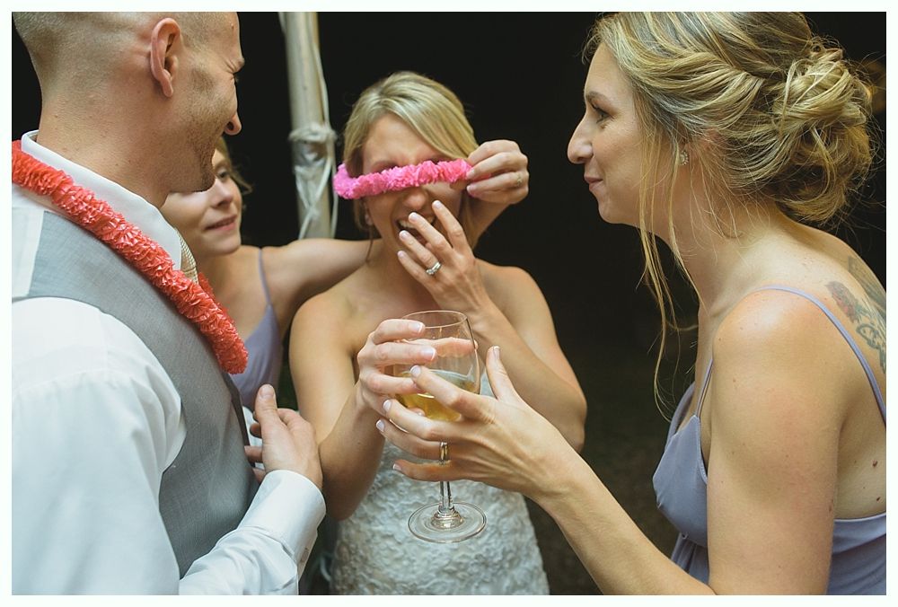 People at a wedding; woman being blindfolded with pink lei while holding a drink.