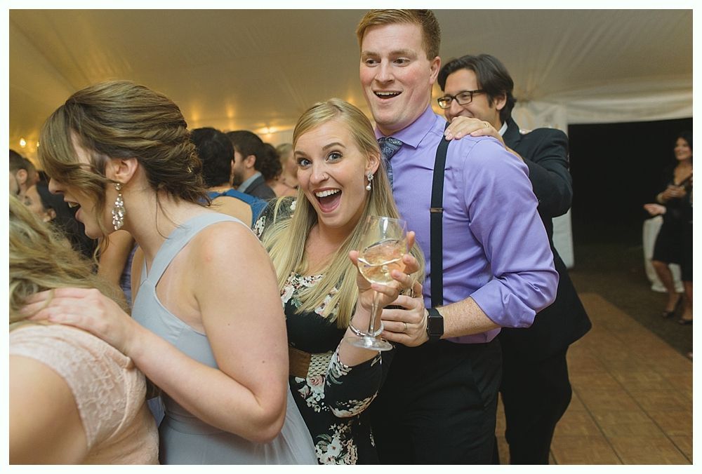 People dancing at a wedding reception, woman holding a drink smiling, man with suspenders smiling.