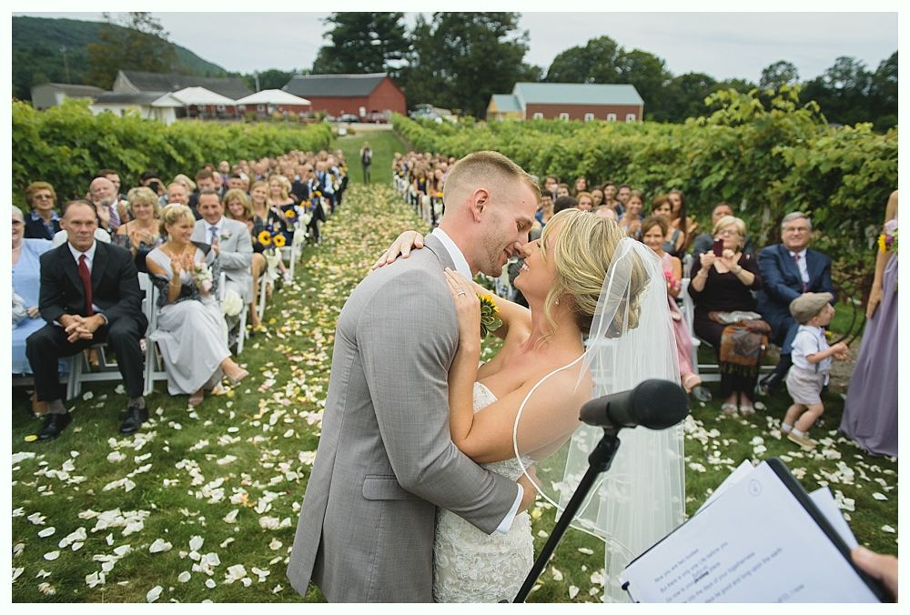 Bride and groom kissing at a wedding ceremony in a vineyard, surrounded by guests.
