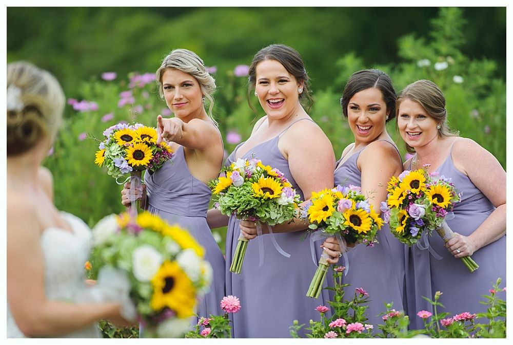 Bridesmaids in lavender dresses holding sunflower bouquets laugh, pointing at bride in a flower field.