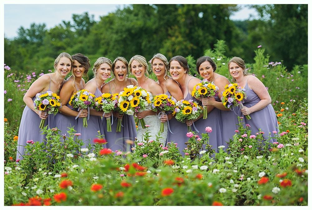 Bridesmaids in lilac dresses holding sunflower bouquets, smiling in a flower field.