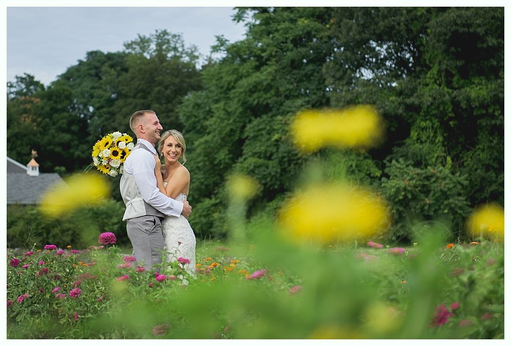 Couple embracing in a field of flowers, smiling; bouquet of sunflowers, trees in the background.
