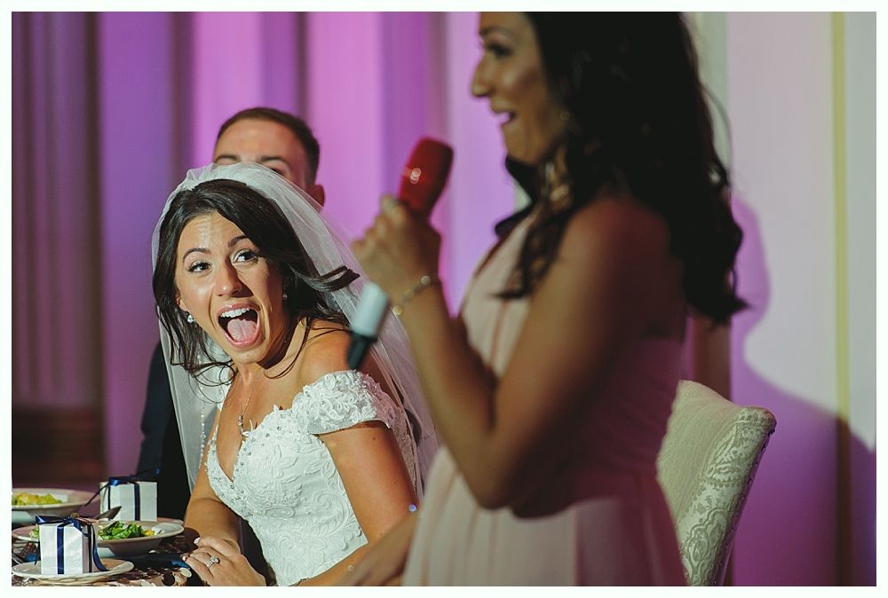 Bride reacts with surprise as a bridesmaid speaks during a wedding reception.