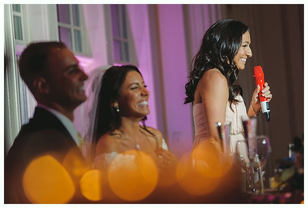 Woman speaking into a red microphone at a wedding reception; bride and man seated, smiling. Purple lighting.