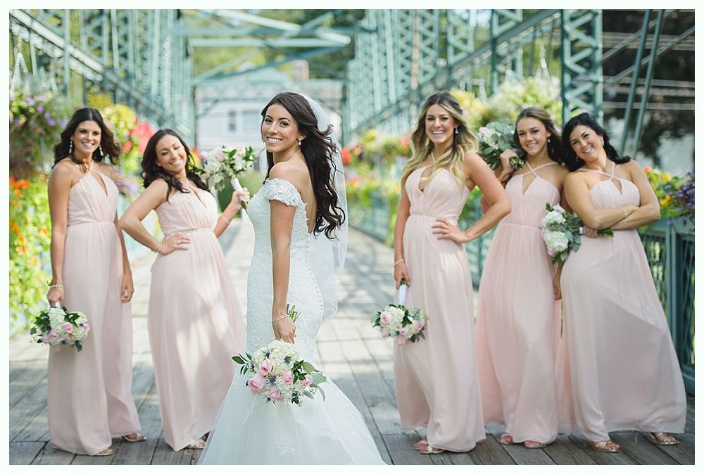 Bride and bridesmaids in pink dresses pose on a bridge with flower baskets.