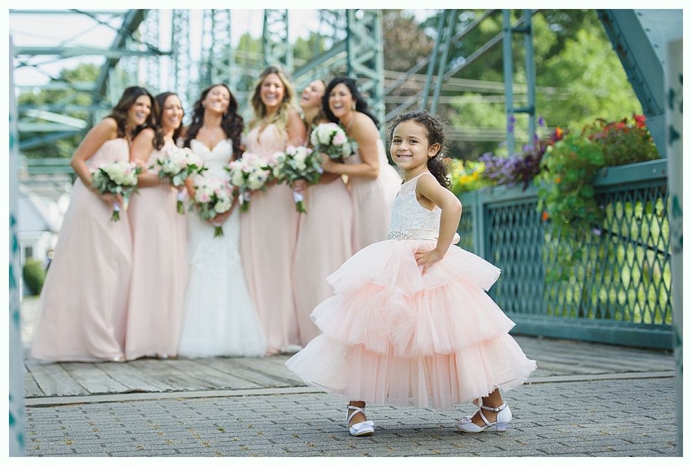 Flower girl in pink dress poses on bridge with laughing bridesmaids in blush gowns.