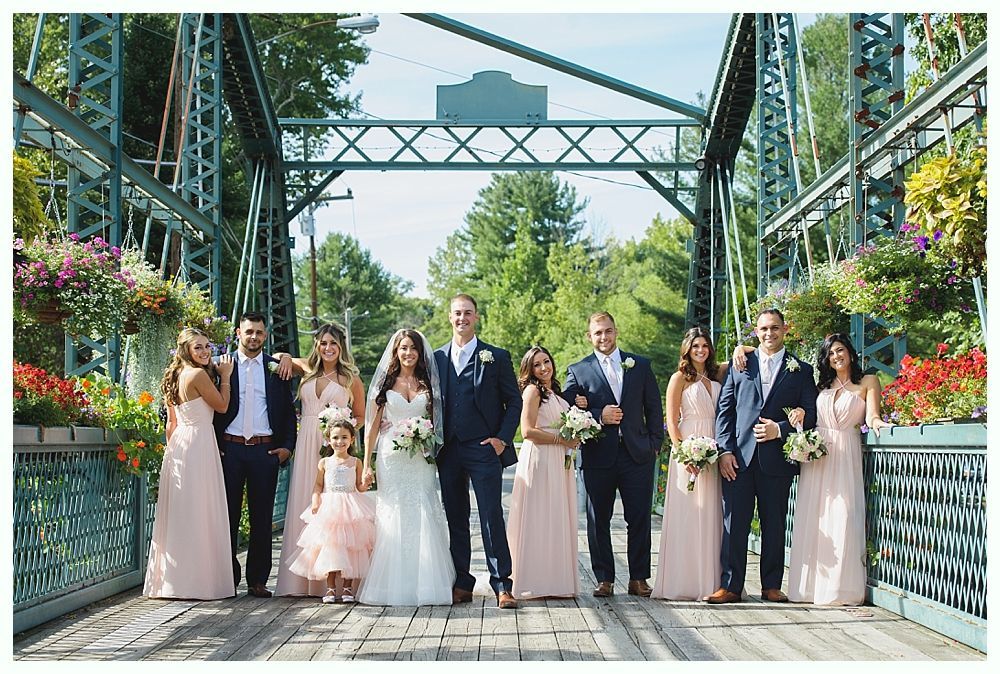 Wedding party on a bridge: bride and groom center, bridesmaids in pink, groomsmen in navy suits, bright floral arrangements.
