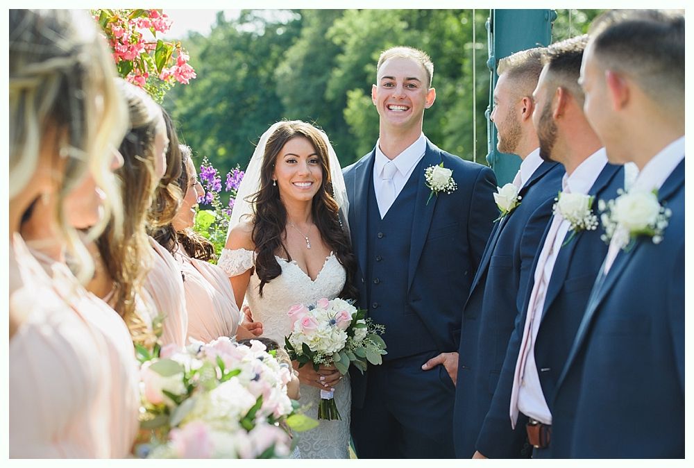 Bride and groom smile, surrounded by bridesmaids and groomsmen, during an outdoor wedding.