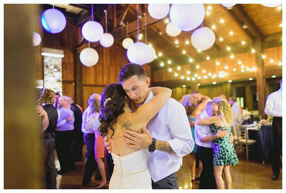 Couple slow dancing at a wedding reception; string lights, paper lanterns, and guests in the background.