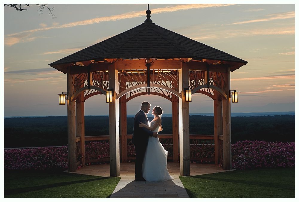 Couple embraces under a gazebo at sunset. Bride in white dress, groom in suit.