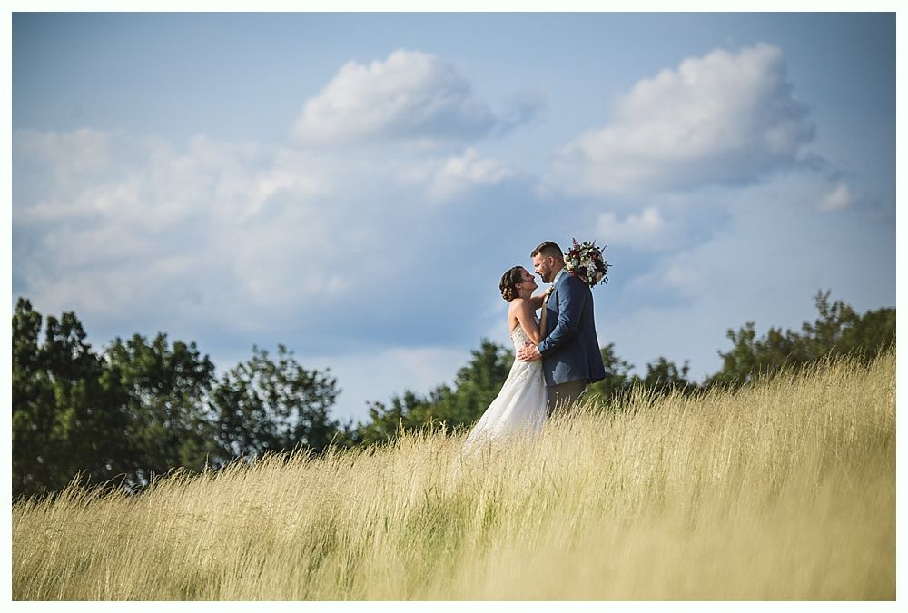 Bride and groom embracing in a grassy field, under a cloudy sky.