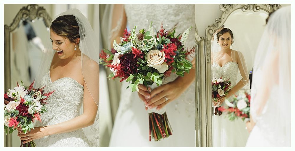 Bride in a white gown and veil holding a bouquet, looking at herself in a mirror and smiling.