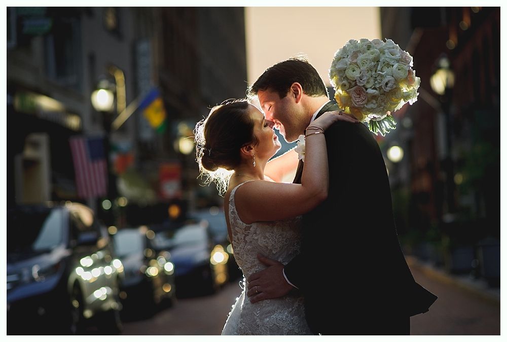 Bride and groom embracing, lit by sunlight, holding bouquet, city street background.