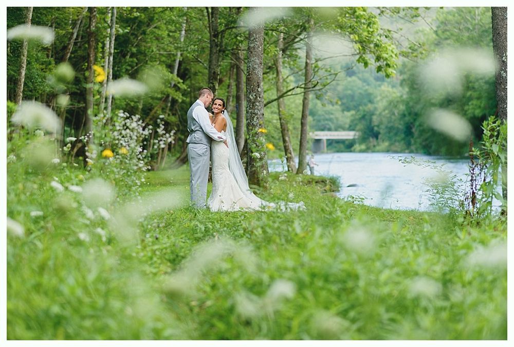 Couple embraces near a river, surrounded by greenery. Wedding attire, serene setting.