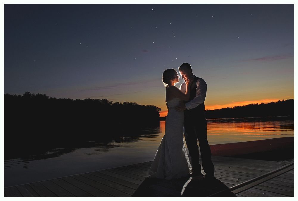 Couple embracing on a dock at dusk, illuminated by a warm glow with water and trees in the background.