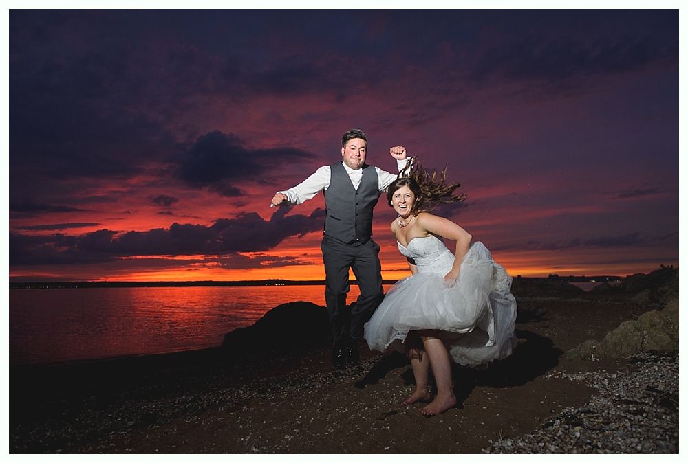 Couple jumping for joy at sunset over water. Bride in white dress, groom in vest.