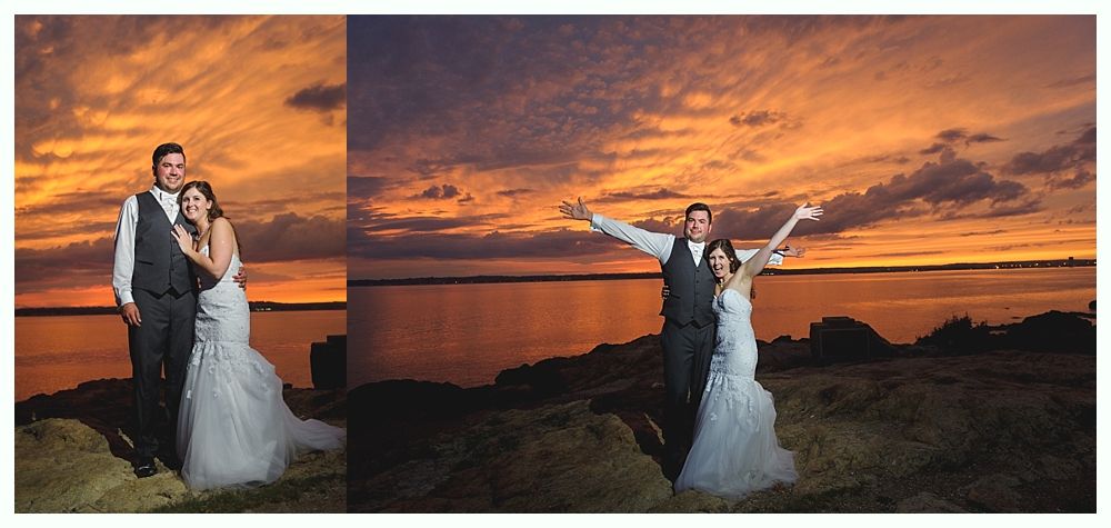 Wedding couple posing on a rocky shore at sunset, arms outstretched, with a fiery sky and calm water.