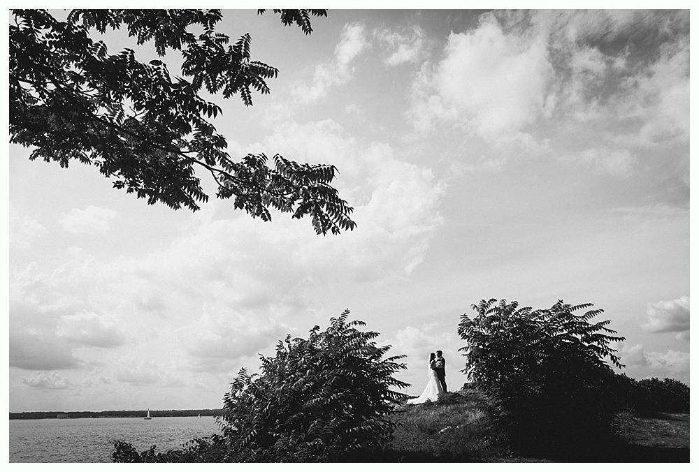 Couple stands on a hillside overlooking water, framed by trees and a cloudy sky. Black and white.