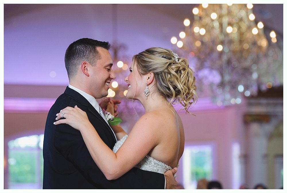 Couple dancing at a wedding reception, smiling at each other under a chandelier.