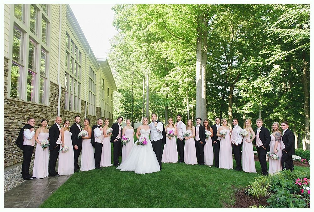 Wedding party poses outdoors in front of a building and trees; bride and groom centered.
