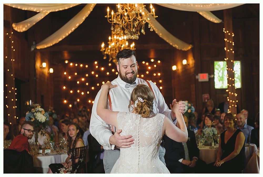 Bride and groom dance at reception; soft lighting, chandelier, and guests at tables.