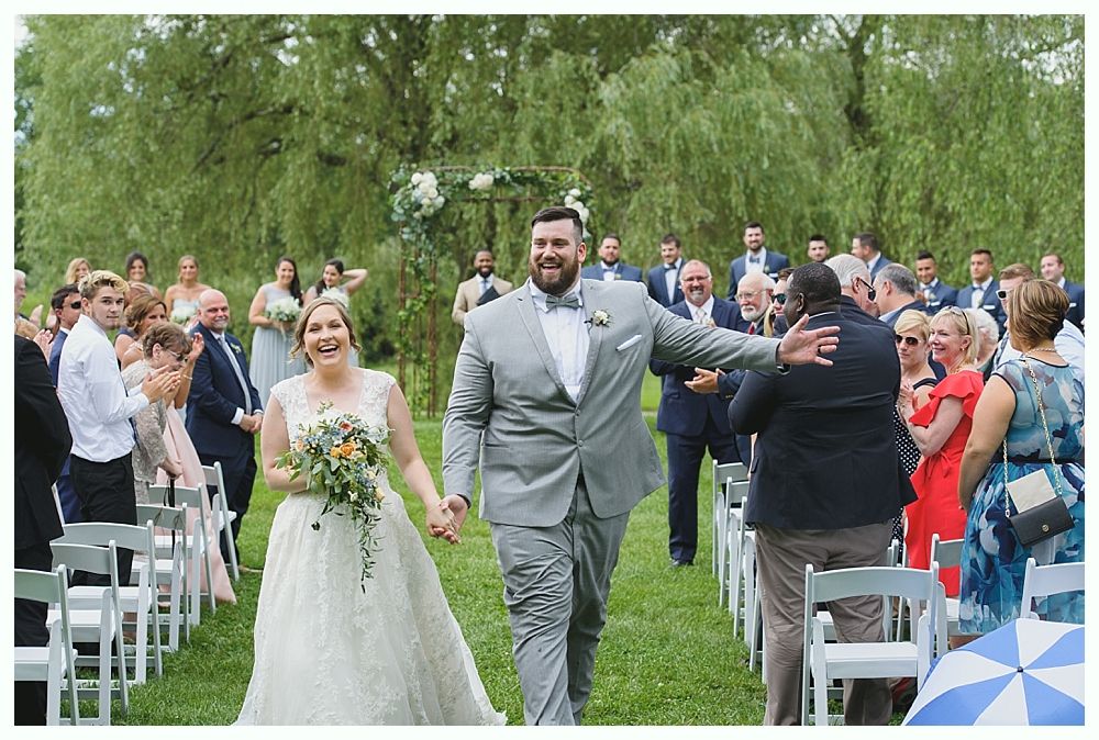 Newlyweds walking down an aisle, holding hands, smiling, amidst wedding guests, outdoor setting, willow trees.