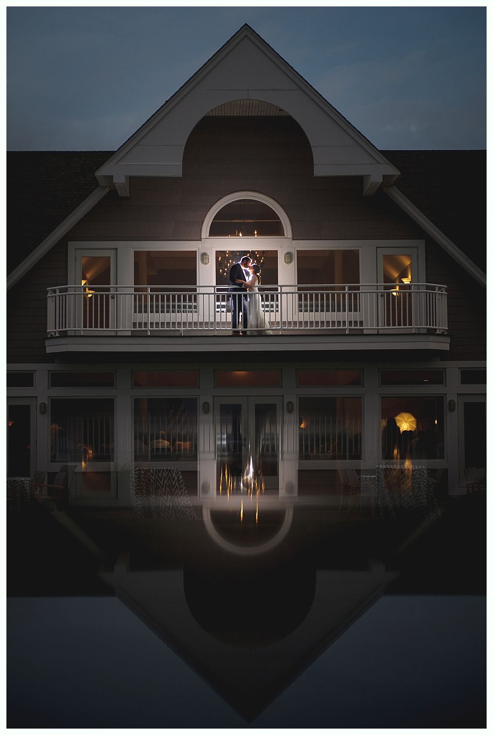 Couple embraces on a balcony at night, reflected in calm water below a building with lights.