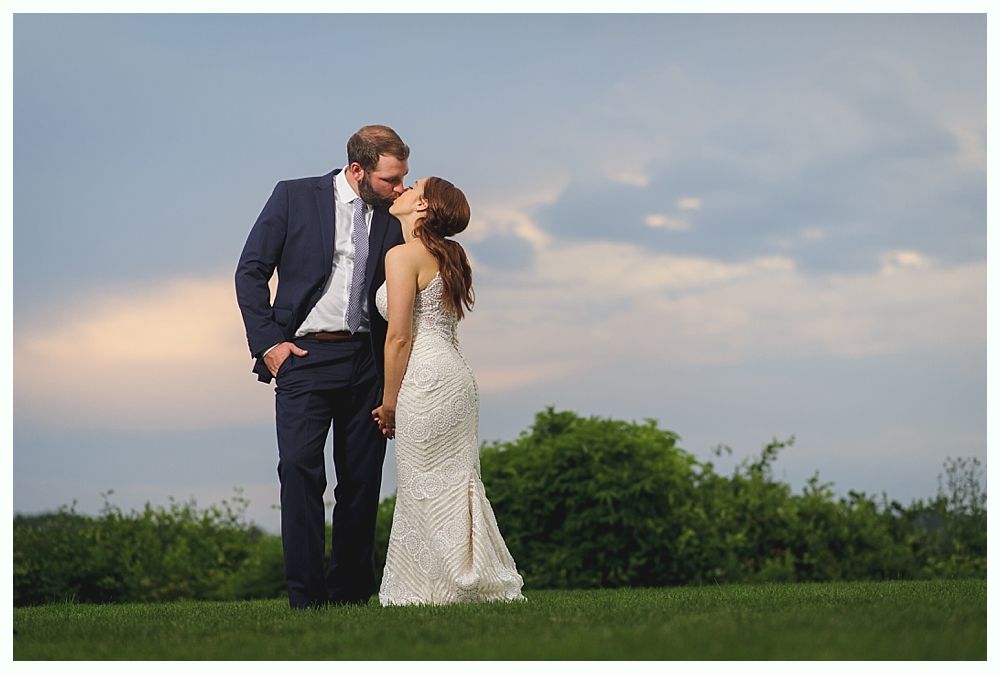 Couple kissing outdoors on a grassy hill; man in a suit, woman in a white wedding dress, cloudy sky.