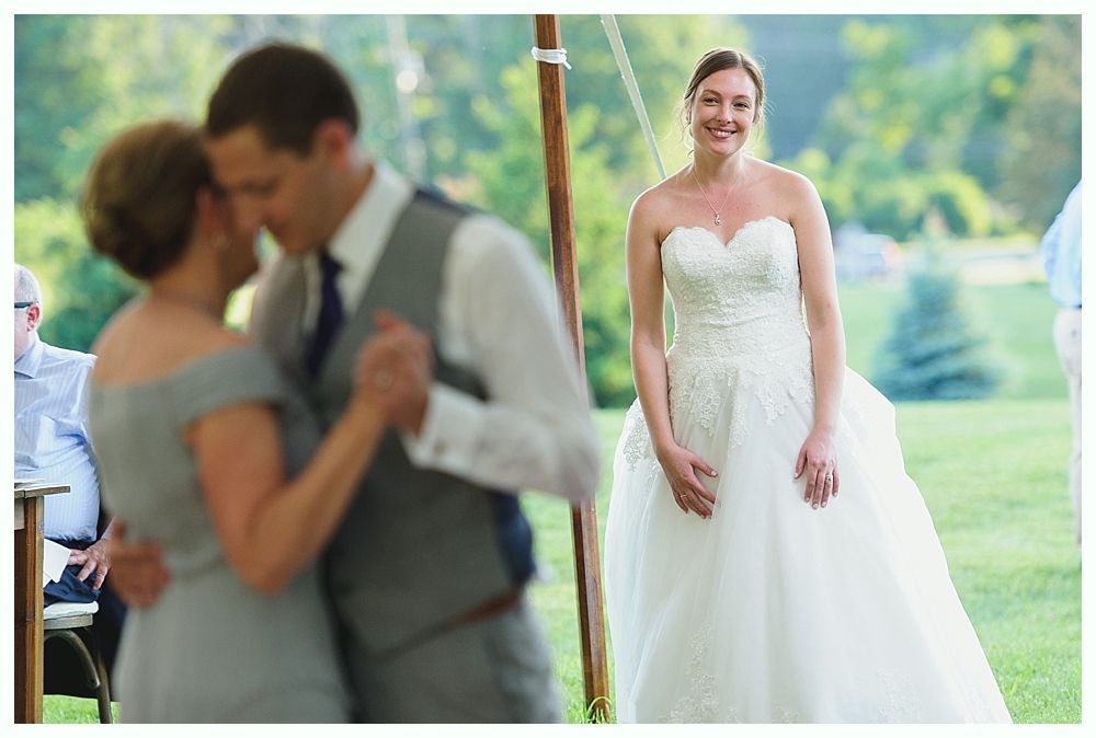 Bride in white strapless gown smiles, watching a couple dancing outdoors at a wedding.