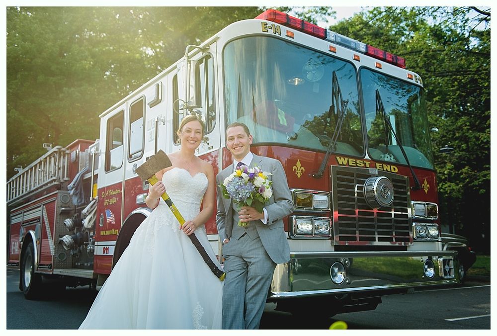 Bride and groom pose with a fire truck. The bride holds an axe, the groom flowers. Outdoors.