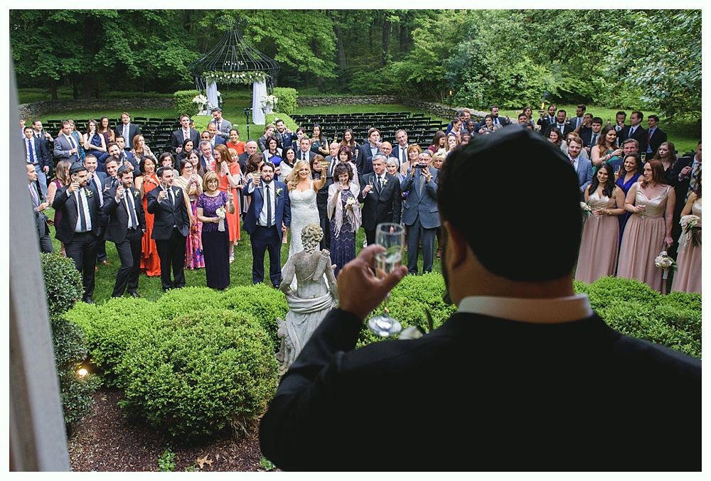 Man toasting with a glass, looking at wedding guests gathered outdoors, trees in the background.
