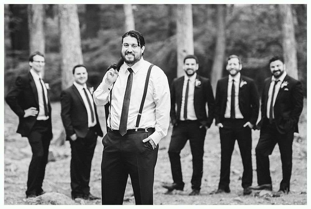 Groom and groomsmen in black suits pose outdoors near trees. Groom holds jacket, smiles.