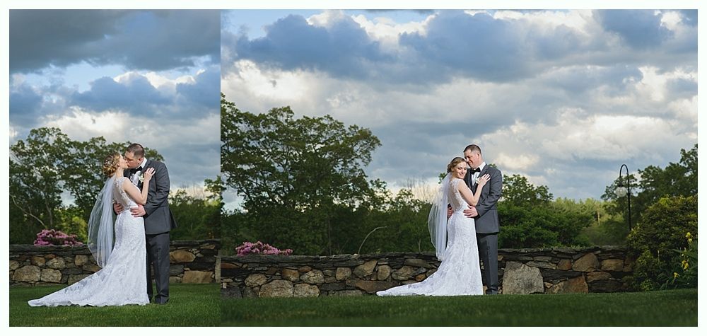 Newlyweds embrace outdoors under a cloudy sky; bride in white dress, groom in gray suit.