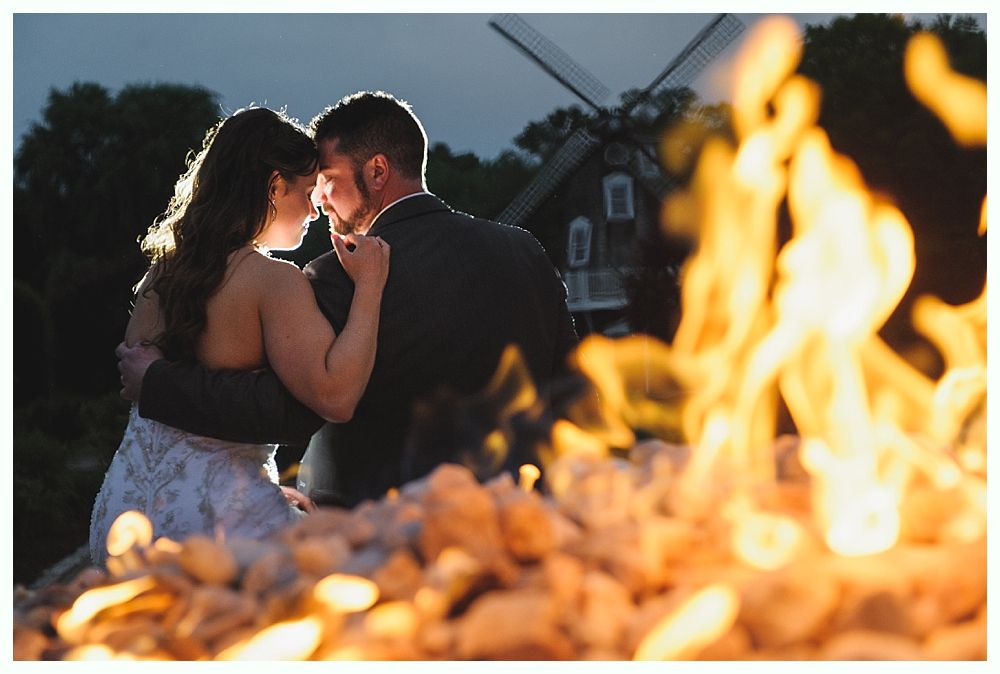 Couple embraces, backlit by a fire, near a windmill at dusk.